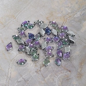 May include: A collection of iridescent glass beads in shades of purple, green, and blue. The beads are faceted and have a diamond shape. They are arranged in a loose pile on a light grey surface.