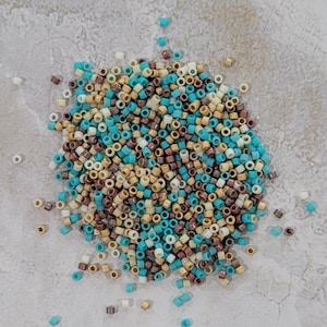 Könnte beinhalten: Ein Haufen kleiner, zylindrischer Perlen in verschiedenen Farben, darunter Türkis, Braun, Beige und Weiß. Die Perlen sind auf einer hellgrauen Oberfläche verstreut. Diese Perlen eignen sich für die Schmuckherstellung und für Bastelarbeiten.