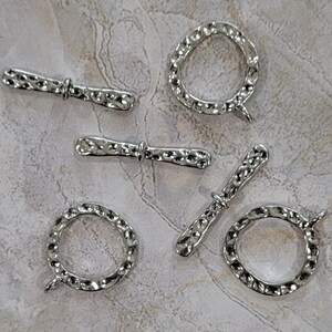 May include: Silver-toned toggle clasps and rings with a hammered texture. The clasps are bar-shaped with a central ring, and the rings have a circular design. These jewelry findings are suitable for necklaces and bracelets.