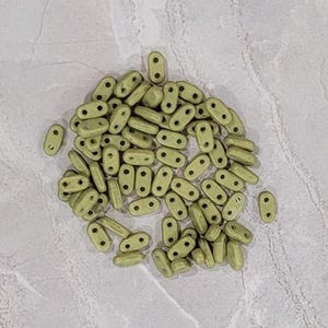 May include: A pile of small, light green oval-shaped beads with two holes in each bead.