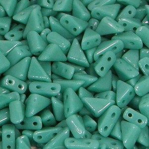 May include: A collection of turquoise-colored glass beads. The beads are a mix of triangular and rectangular shapes, each with two holes for stringing. The beads have a glossy finish and are scattered, creating a textured appearance.