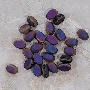 Puede incluir: Una colección de cuentas de vidrio ovaladas iridiscentes en tonos de morado y negro. Las cuentas tienen una superficie texturizada y están dispersas sobre un fondo gris claro.