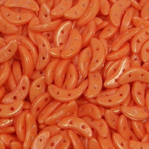 May include: A close-up of a large quantity of small, orange, crescent-shaped glass beads. The beads have a pearlescent finish and are arranged in a random pattern.