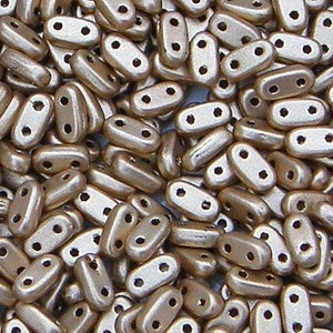 May include: A collection of small, oblong, metallic gold-colored beads, each featuring two distinct holes positioned side-by-side. These uniform, pill-shaped beads have a smooth, reflective surface and are arranged in a dense, overlapping pile.