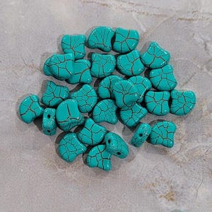 May include: A collection of turquoise-colored, heart-shaped beads with a cracked, dark brown pattern. These beads are scattered on a light gray, textured surface. The beads have holes for stringing, suggesting they are for jewelry making.