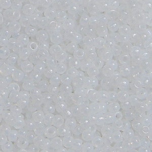 Puede incluir: Primer plano de un montón de pequeñas cuentas de semillas blancas translúcidas. Las cuentas son redondas con un pequeño agujero en el centro. Las cuentas están dispersas, creando una superficie texturizada.