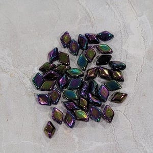 Op de afbeelding: Een verzameling iriserende, ruitvormige kralen. De kralen vertonen een kleurverschuivingseffect, met tinten paars, groen en goud. Ze zijn verspreid over een lichtgrijze, getextureerde ondergrond, wat wijst op een knutsel- of sieraden thema.