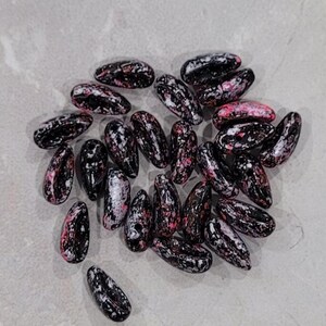 May include: A collection of black and pink speckled beans, with white accents, arranged in a loose pile on a light gray surface.