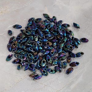 Op de afbeelding: Een stapel iriserende blauwe en paarse glazen kralen. De kralen zijn klein en ovaalvormig met een gat in het midden.