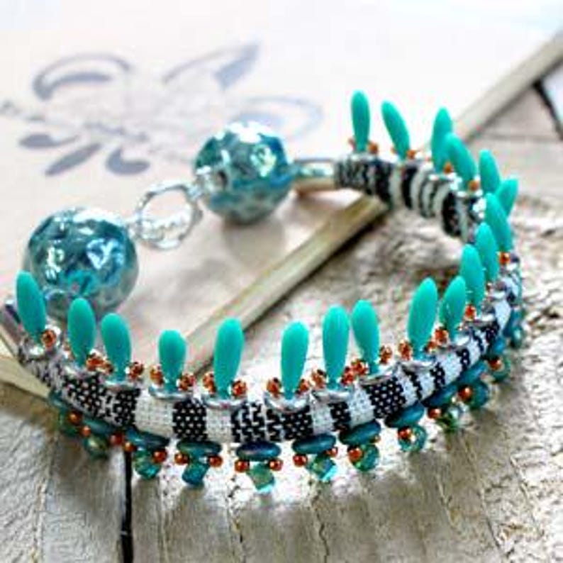 May include: A turquoise and black beaded bracelet with a silver clasp and a turquoise bead charm. The bracelet is made with a woven fabric and has a fringe of turquoise beads.