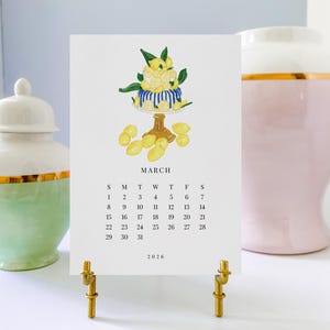 May include: A March calendar page with a lemon-themed illustration, displayed on a gold stand. The calendar features a lemon cake design and the year 2020. Decorative vases in green and pink flank the calendar.