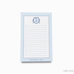 May include: A light blue notepad with a white interior and a blue border. The top features a crest with the letter "M". The notepad has a checklist format with lines and check boxes. The text "THE MITCHELL FAMILY" is at the bottom.