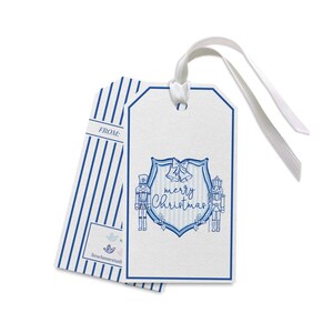 Könnte beinhalten: Weiße Weihnachtsgeschenkanhänger mit blauen Akzenten und einem weißen Band. Ein Anhänger zeigt ein blaues Schilddesign mit den Worten "merry Christmas" und Nussknackern. Der andere hat blaue vertikale Streifen.