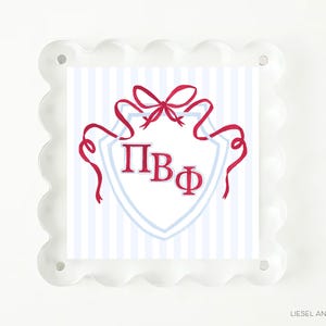 Puede incluir: Un marco de acrílico transparente con borde festoneado contiene una impresión con un fondo a rayas azul claro y blanco. La impresión presenta un diseño de lazo y cinta rojos con las letras griegas ΠΒΦ en rojo.