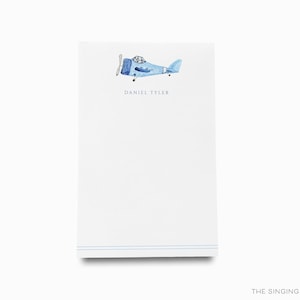 Op de afbeelding: Gepersonaliseerd notitieblok met een blauwe vliegtuigillustratie en de naam "Daniel Tyler" gedrukt in paarse tekst.