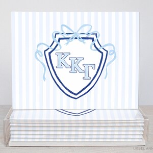May include: A set of stationery cards with a light blue and white striped pattern. Each card displays a shield design with the letters KKT in light blue, framed by a dark blue border and ribbons. The cards are presented in a clear acrylic holder.