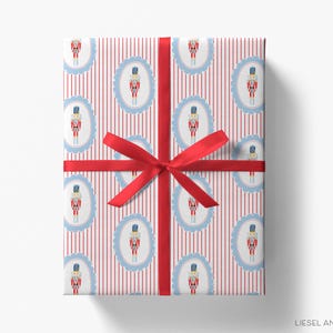 Puede incluir: Regalo envuelto con cinta roja. El papel de regalo presenta un patrón de ilustraciones de cascanueces dentro de marcos ovalados azules, sobre un fondo de rayas verticales rojas y blancas. El lazo es de color rojo brillante.