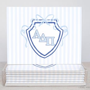 May include: Stationery cards with a light blue and white striped pattern. Each card displays a blue shield design with the Greek letters A, Δ, and Π. The shield is decorated with a bow and ribbons.