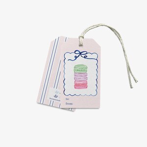 Puede incluir: Dos etiquetas de regalo rectangulares con fondo rosa claro. La etiqueta frontal presenta una ilustración en acuarela de una pila de tres macarons en verde, lavanda y rosa, enmarcada por un borde festoneado azul con un lazo. Incluye to: y from:.