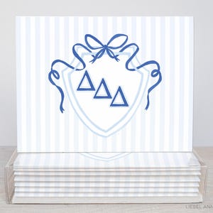 May include: Stationery cards with a blue and white striped pattern, featuring a blue crest design with three triangles and a bow. The cards are stacked in a clear acrylic holder.