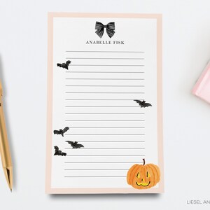 a pink sticky note pad with a black bat illustration, a gold pen, and a small pumpkin decoration.