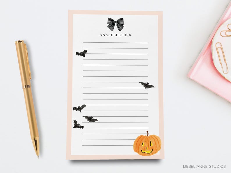 a pink sticky note pad with a black bat illustration, a gold pen, and a small pumpkin decoration.