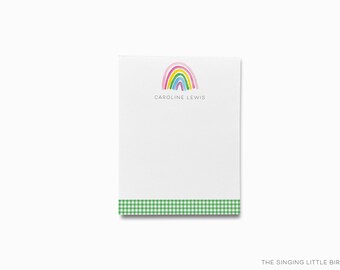 May include: A white notepad with a watercolor rainbow design at the top and the name "CAROLINE LEWIS" printed below it. The bottom edge features a green and white gingham pattern. The notepad is on a white background.