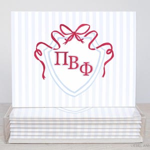 May include: A set of stationery cards with a shield design. The cards have a light blue and white striped background. The shield is outlined in light blue with red accents, including a bow and ribbons. The letters "ΠΒΦ" are in red.