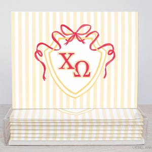 May include: A set of stationery cards with a cream and yellow striped pattern. Each card displays a shield design with red ribbons and the Greek letters X and Ω. The cards are presented in a clear acrylic box.