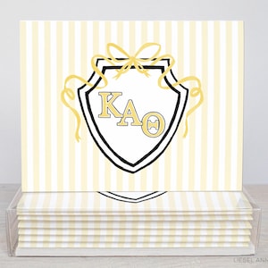 May include: A stationery set with a shield design, featuring the letters KAO in gold, and gold bow and ribbons. The background has vertical yellow and white stripes. The set is presented in a clear acrylic box.