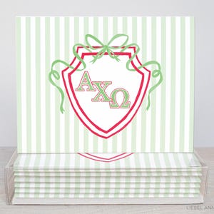 May include: A set of stationery cards with a green and white striped pattern. Each card displays a red and green shield emblem with the Greek letters A, X, and Ω. The cards are presented in a clear plastic box.