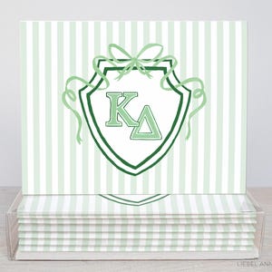 May include: A stack of stationery cards with a green and white striped background. Each card features a green shield design with the letters K and Δ. The shield is adorned with a bow and ribbons.