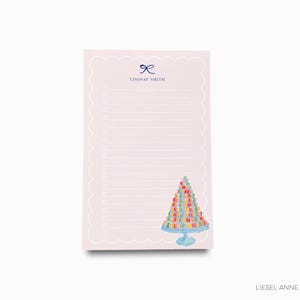 May include: Pink notepad with a scalloped edge and a white lined interior. The notepad has a blue ribbon bow and the text "Lindsay Smith" printed at the top. The bottom of the notepad features a watercolor illustration of a tower of colorful macarons on a blue pedestal.