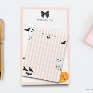 a pink and white striped notepad with a Halloween-themed design, including a ghost, bats, and a pumpkin. There is also a gold pen and a pink object on the right side of the image.