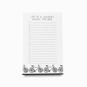 May include: White notepad with black lines and a black and white illustration of bicycles at the bottom. The notepad has the text "LIFE IS A JOURNEY ENJOY THE RIDE" at the top.