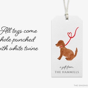 May include: A brown dog with a red bow tie is holding a red ribbon with a heart shape at the end. The text "a gift from, THE HAMMILLS" is below the dog. The text "All tags come hole punched with white twine" is on the left side of the image.