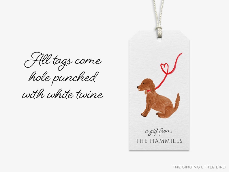 May include: A brown dog with a red bow tie is holding a red ribbon with a heart shape at the end. The text "a gift from, THE HAMMILLS" is below the dog. The text "All tags come hole punched with white twine" is on the left side of the image.