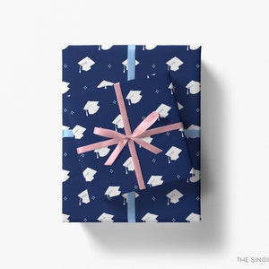 May include: Two navy blue gift boxes with a white graduation cap pattern. The smaller box is wrapped with a pink ribbon and the larger box is wrapped with a light blue ribbon.