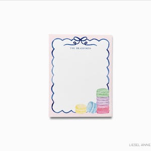 May include: A pink notepad with a blue scalloped border and a blue bow. The notepad has the text "The Bradfords" at the top. There are five colorful macarons in the bottom right corner.