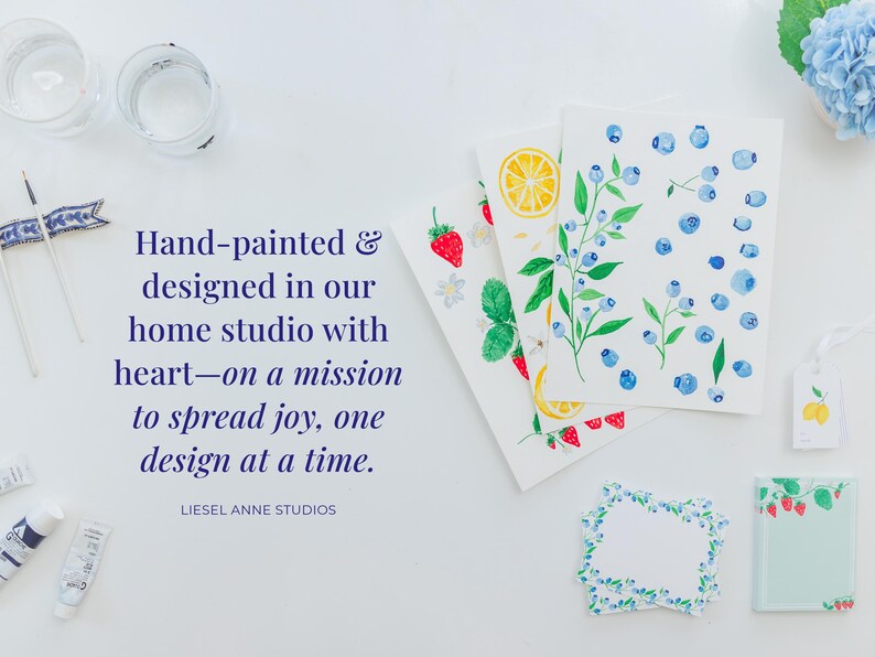 a hand-painted and designed home studio with a quote from Liesel Anne Studios about spreading joy through one design at a time. The studio has various art supplies and decorative items, including watercolor paintings, a blueberry design, and a blueberry-themed frame.