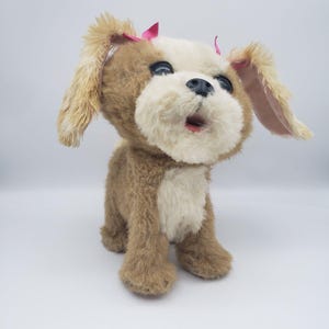 Op de afbeelding: Een pluchen hond met bruine en witte vacht, grote zwarte ogen en een roze strik. De mond van de hond staat open, alsof hij blaft of zingt. Het speelgoed is ontworpen om op een puppy te lijken.