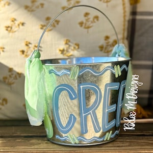 May include: A galvanized metal bucket with a silver handle and light green fabric accents. The word "CREE" is painted in blue with a white outline. The bucket has a wavy blue and green design.
