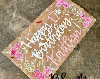 Puede incluir: Un letrero rectangular de papel marrón con las palabras "Happy 17th Birthday Kaitlyn!" en escritura blanca y rosa. El letrero está decorado con rosas rosas y hojas verdes. La esquina inferior derecha tiene el texto "Ridee M Designs".
