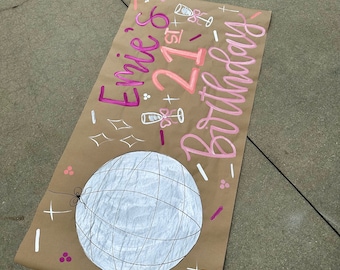 May include: A brown paper banner with pink and white lettering that says "Emie's 21st Birthday". The banner also has a white disco ball drawn on it and pink and white confetti.