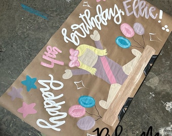 Puede incluir: Una pancarta de papel marrón con las palabras "Happy birthday Ellie!" escritas en pintura blanca. Una niña con un leotardo rosa está pintada en la pancarta, haciendo una parada de manos en una viga de equilibrio. Hay estrellas y globos rosas, azules y turquesa pintados en la pancarta. Las palabras "Ridlee M. Designs" están escritas en pintura negra en la parte inferior de la pancarta.