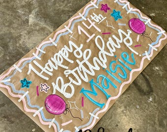 Puede incluir: Papel marrón con la frase "Happy 14th Birthday Maisie" en escritura blanca y azul. El diseño incluye líneas onduladas rosas y azules, estrellas y globos rosas. La esquina inferior derecha tiene el texto "Ridee M Designs".