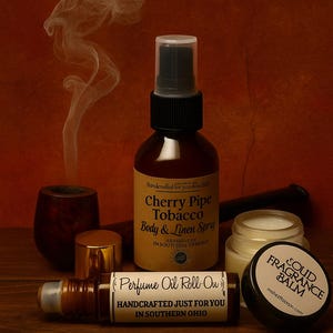 May include: A brown glass spray bottle labeled Cherry Pipe Tobacco Body & Linen Spray, a perfume oil roll-on, a small jar of solid fragrance balm, and a dark wooden pipe with smoke rising, all arranged together on a dark wooden surface.