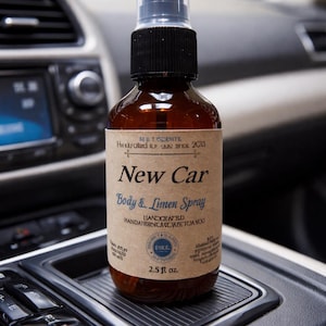 May include: A brown glass spray bottle labeled "New Car Body & Linen Spray" with a black cap and clear spray nozzle. The bottle is 2.5 fl oz and sits in a car's console. The label includes the text "Handcrafted" and other text.
