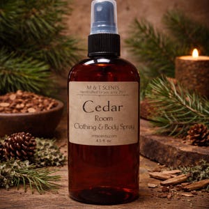 May include: A brown glass spray bottle labeled "Cedar Room Clothing & Body Spray" with a black cap. The label reads "M & T SCENTS Handcrafted for you since 2013" and "4.5 fl. oz." The background includes pine needles, a lit candle, and a wooden bowl.