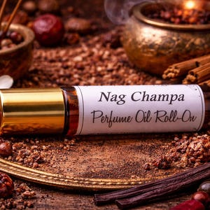 May include: A cylindrical glass perfume oil roll-on bottle with a gold-colored cap. The white label features the black printed text Nag Champa Perfume Oil Roll-On. The bottle is positioned horizontally, showcasing its clear glass body and metallic lid.